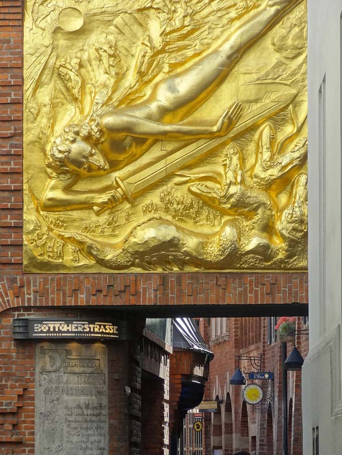 A large bronze relief shows a naked young man fighting with a sword against a three-headed being. It adorns the entrance to Böttcherstrasse, which can be seen in the background.