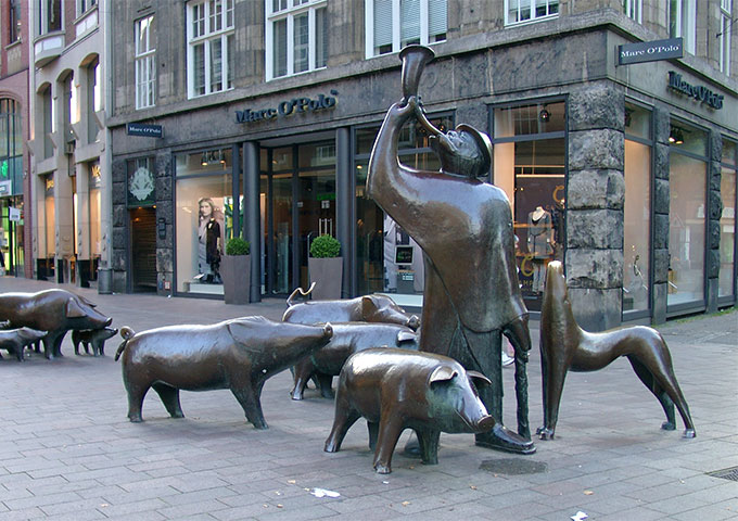 Seitenansicht des Denkmals "Hirt mit Schweinen". Es zeigt den Hirten aus Bronze mit seinem Hund sowie den Schweinen. In seinen Händen trägt er ein Horn und einen Stock.