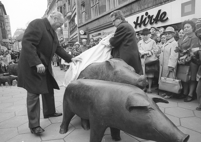 Zwei Herren enthüllen das Denkmal "Hirt mit Schweinen" in der Sögestraße. Passanten stehen im Hintergrund und beobachten das Geschehen.